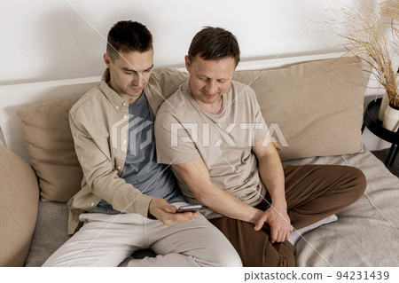 Happy gay couple with casual clothes spending time together at home and watching something on smartphone, or having video call with friends. Homosexual relationships and alternative love. 94231439