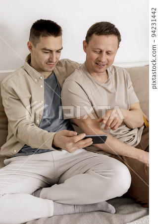 Happy gay couple with casual clothes spending time together at home and watching something on smartphone, or having video call with friends. Homosexual relationships and alternative love. 94231442