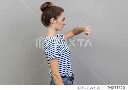 Side view of unhappy unsatisfied young adult woman wearing striped T-shirt showing thumb down, dislike gesture, negative emotions, bad rating. Indoor studio shot isolated on gray background. 94231620