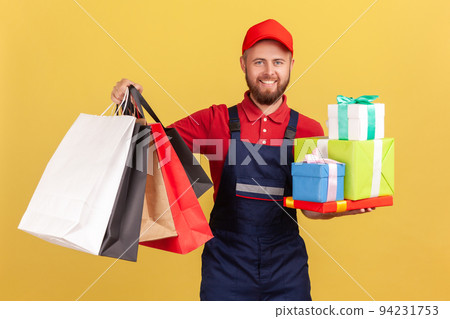 Portrait of smiling positive optimistic bearded courier man standing with many present boxes and shopping bags, door to door delivery. Indoor studio shot isolated on yellow background. 94231753