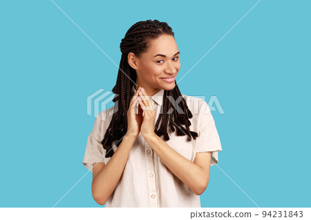 Portrait of thoughtful woman with black dreadlocks schemes something, keeps fingers together, considers over cunning plans, wearing white shirt. Indoor studio shot isolated on blue background. Portrait of thoughtful woman with black dreadlocks schemes something, keeps fingers together, considers over cunning plans, wearing white shirt. Indoor studio shot isolated on blue background. 94231843