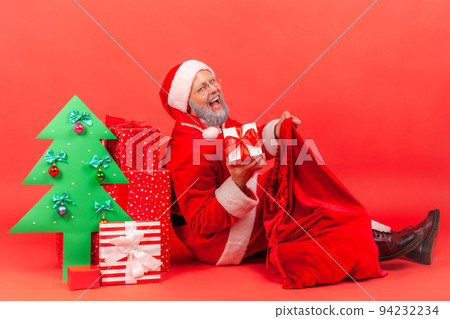 Happy positive elderly man with gray beard wearing santa claus costume with festive mood, taking present box from bag, looking at camera and smiling. Indoor studio shot isolated on red background. 94232234
