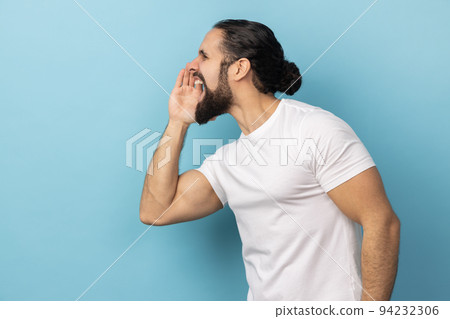 Attention, announcement. Side view of handsome man with beard wearing white T-shirt screaming message, loud advertising, keeps hands near mouth. Indoor studio shot isolated on blue background. 94232306