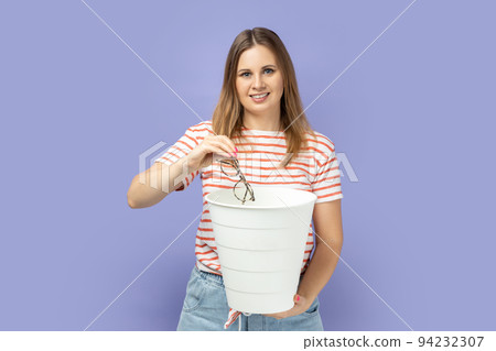 Portrait of blond woman wearing striped throwing out her optical glasses after laser vision treatment, being happy to have better eyesight. T-shirt Indoor studio shot isolated on purple background. 94232307