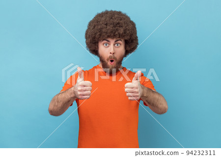 Shocked man with Afro hairstyle wearing orange T-shirt showing thumbs up, looking at camera with open mouth, astonishing with excellent result. Indoor studio shot isolated on blue background. 94232311