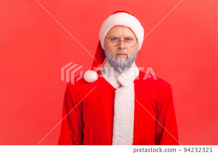 Portrait of serious elderly man with gray beard wearing santa claus costume looking at camera with strict and bossy facial expression. Indoor studio shot isolated on red background. 94232321