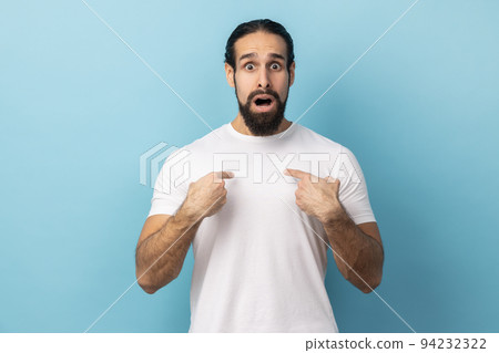 Portrait of amazed happy excited man with beard wearing white T-shirt pointing himself, feeling proud and ambitious, shocked by huge success. Indoor studio shot isolated on blue background. 94232322