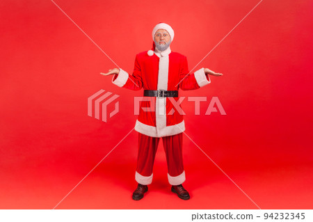 Full length portrait of elderly man with gray beard wearing santa claus costume shrugging shoulders as doesn't know answer, being uncertain, not sure. Indoor studio shot isolated on red background. 94232345
