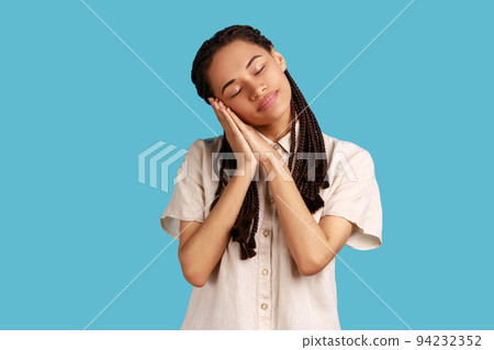 Portrait of beautiful woman with black dreadlocks sleeping laying down on her palms, having comfortable nap and resting, dozing off, wearing white shirt. Indoor studio shot isolated on blue background 94232352