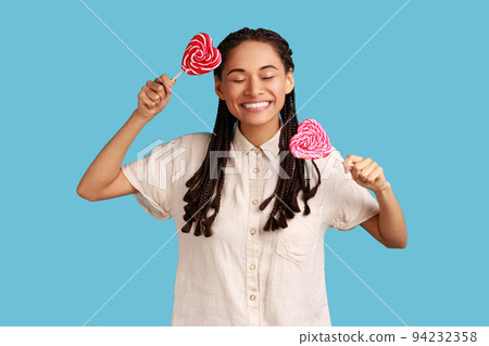 Excited woman woman with black dreadlocks holding two candies with heart shape, keeps eyes closed, has happy positive expression, wearing white shirt. Indoor studio shot isolated on blue background. 94232358