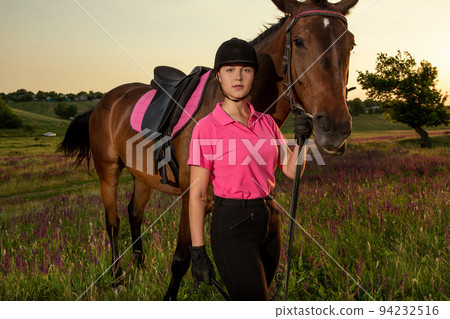 Beautiful smiling girl jockey stand next to her brown horse wearing special uniform on a sky and green field background on a sunset. Beautiful smiling girl jockey stand next to her brown horse wearing special uniform on a sky and green field background on a sunset. 94232516
