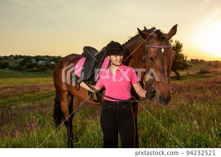 Beautiful smiling girl jockey stand next to her brown horse wearing special uniform on a sky and green field background on a sunset. 94232521
