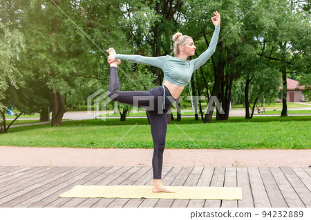 A slender woman on a wooden platform in summer, does flow yoga in park 94232889