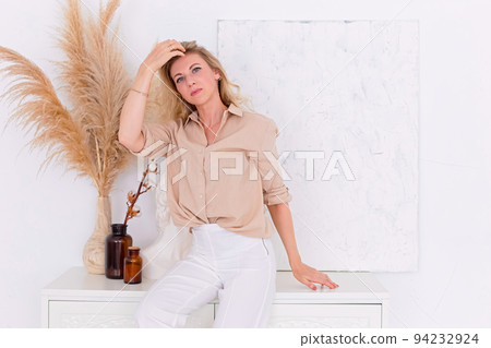 A slim woman is standing in the interior of a white room, near the wall 94232924