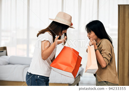 Two Asian young women holding shopping paper bag looking talking together in bedroom 94233355