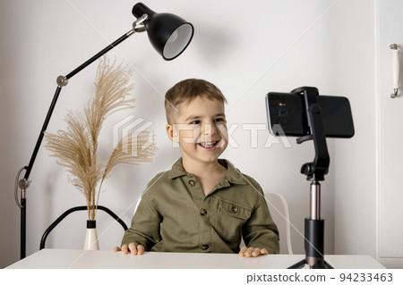 Adorable, cute, little boy blogger recording lifestyle blog, talking to camera of smartphone on tripod. Young influencer filming vlog for his channel. Child makes video for his followers online. Adorable, cute, little boy blogger recording lifestyle blog, talking to camera of smartphone on tripod. Young influencer filming vlog for his channel. Child makes video for his followers online. 94233463