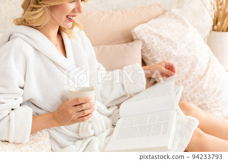 Young woman in white terry robe is drinking coffee and reading magazine or book in bedroom. Young woman in white terry robe is drinking coffee and reading magazine or book in bedroom. 94233793