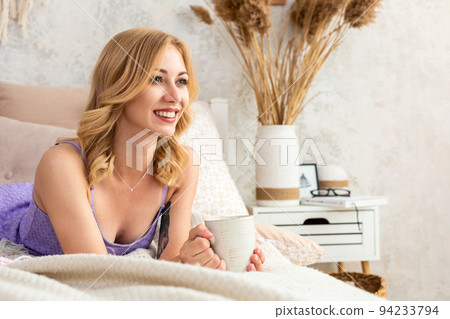 Young woman holding a mug with tea or coffee sitting on the bed in the bedroom. Near lie yarn and knitting needles. Young woman holding a mug with tea or coffee sitting on the bed in the bedroom. Near lie yarn and knitting needles. 94233794
