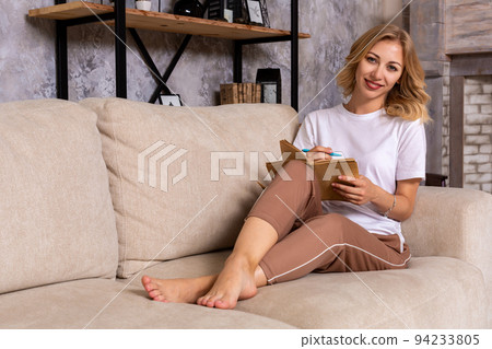 Thoughtful pensive woman sitting on sofa in modern white apartment, writing list to do in notepad using pen. 94233805