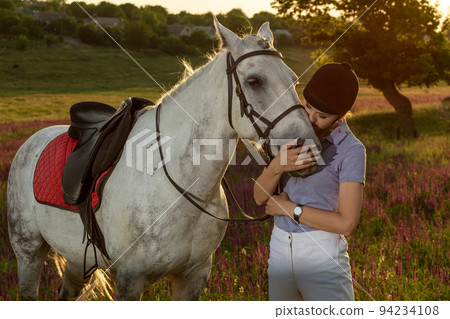 Jockey young girl petting and hugging white horse in evening sunset 94234108