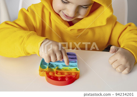 Little boy plays with rainbow Pop it fidget toy. Antistress children game. Children's trendy silicone toys pop it to relieve stress and develop hand motor skills. Colorful hand toy with push bubbles. 94234201
