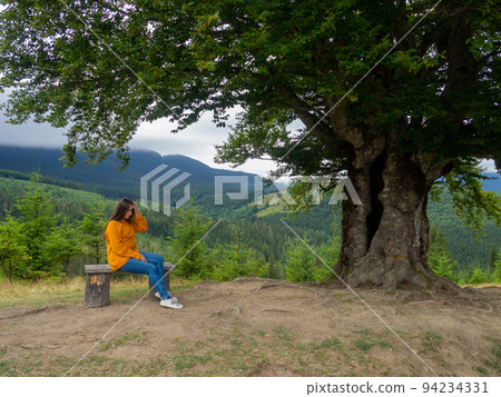 Female in casual clothes sits under large tree with forest background Female in casual clothes sits under large tree with forest background 94234331