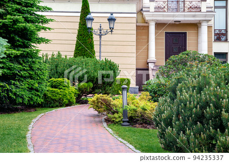 Backyard with bark mulched evergreens pine plants trees and bushes with needles and retro iron twin lantern and ground lighting near brick stone tile pathway at house. Backyard with bark mulched evergreens pine plants trees and bushes with needles and retro iron twin lantern and ground lighting near brick stone tile pathway at house. 94235337