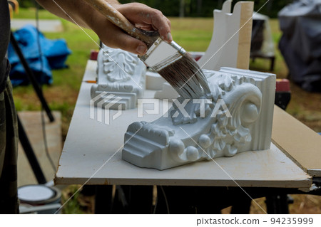 With a paintbrush, a worker paints wooden corbels for a kitchen island on a table surface 94235999