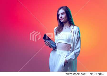 Portrait of young beautiful woman in white casual clothes posing with tablet isolated over red background in neon light Portrait of young beautiful woman in white casual clothes posing with tablet isolated over red background in neon light 94237062
