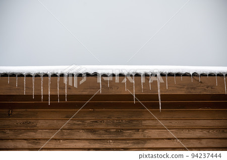 Icicles hanging from  snow-covered roof of house. Cold winter outside the house. 94237444