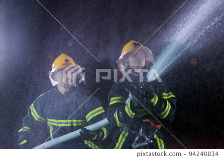 Firefighters using a water hose to eliminate a fire hazard. Team of female and male firemen in dangerous rescue mission. Firefighters using a water hose to eliminate a fire hazard. Team of female and male firemen in dangerous rescue mission. 94240814