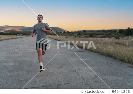 Attractive fit man running fast along countryside road at sunset light, doing jogging workout outdoors 94241268