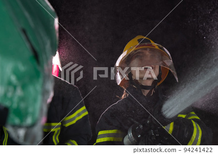 Firefighters using a water hose to eliminate a fire hazard. Team of female and male firemen in dangerous rescue mission.  94241422