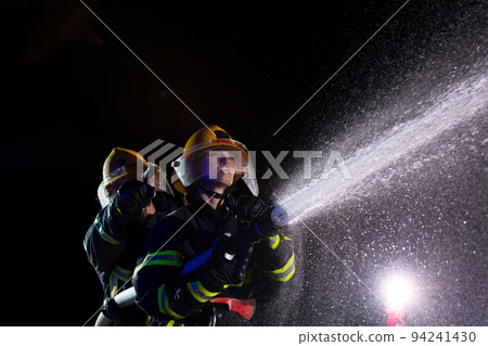 Firefighters using a water hose to eliminate a fire hazard. Team of female and male firemen in dangerous rescue mission. Firefighters using a water hose to eliminate a fire hazard. Team of female and male firemen in dangerous rescue mission. 94241430