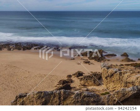 View of empty small sand beach with with long exposure blurred ocean waves and sharp rock and cllifs at wild Rota Vicentina coast near Porto Covo, Portugal. View of empty small sand beach with with long exposure blurred ocean waves and sharp rock and cllifs at wild Rota Vicentina coast near Porto Covo, Portugal. 94242154