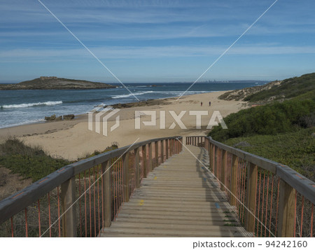 wooden bridge or sidewalk leading to Praia da Ilha do Pessegueiro sand beach with group of hikers, ocean waves and Fort of Pessegueiro small island at Rota Vicentina coast near Porto Covo, Portugal. 94242160