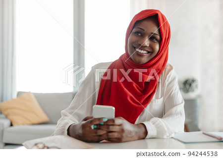 Excited african american muslim businesswoman in hijab using cellphone while sitting at desk at home office 94244538