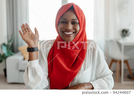 Greeting concept. Portrait of happy black muslim woman waving hello and smiling at camera, using laptop at home office Greeting concept. Portrait of happy black muslim woman waving hello and smiling at camera, using laptop at home office 94244607