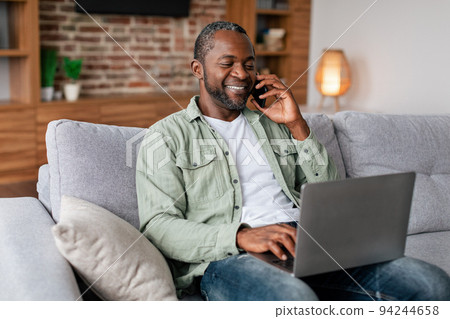 Smiling busy middle aged african american businessman in casual calling by phone and typing on laptop 94244658