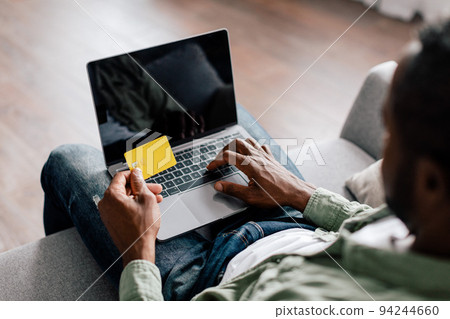 Middle aged african american guy typing on computer with blank screen and showing credit card Middle aged african american guy typing on computer with blank screen and showing credit card 94244660