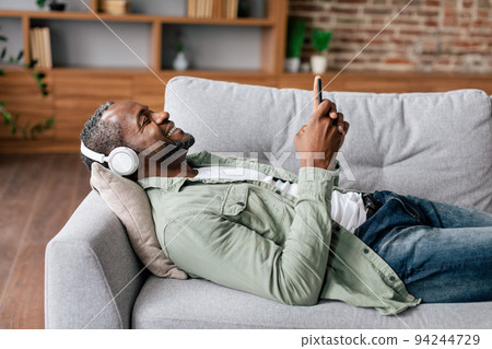 Cheerful mature african american male in casual in wireless headphones enjoys favorite music with smartphone 94244729