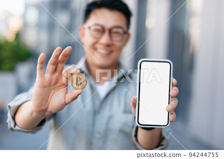 Cryptocurrency app. Asian businessman showing golden bitcoin coin and blank smartphone with white screen, mockup Cryptocurrency app. Asian businessman showing golden bitcoin coin and blank smartphone with white screen, mockup 94244755