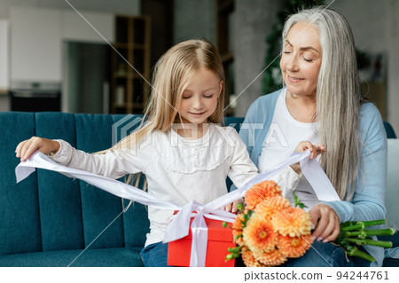 Happy caucasian small granddaughter opens box with gift from elderly grandma with bouquet of flowers 94244761