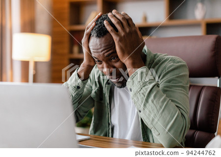 Sad frustrated adult african american guy in casual hold his head with hand watch at laptop, suffer from stress 94244762