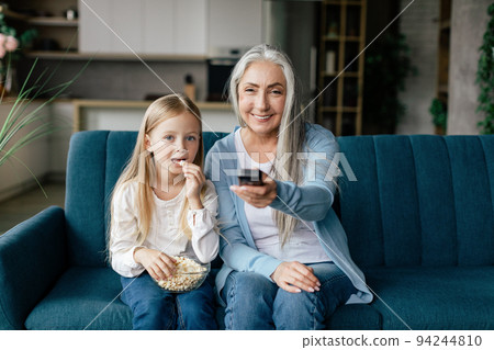Happy caucasian small girl eat popcorn with elderly lady with remote control, watch tv on sofa in living room 94244810