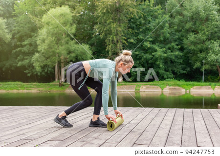 A woman in the summer in the park, unwinds a green gym mat, prepares for sports. A woman in the summer in the park, unwinds a green gym mat, prepares for sports. 94247753