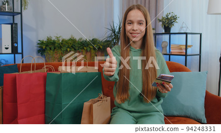 Happy shopaholic young teen girl sitting with shopping bags making online payment with credit card Happy shopaholic young teen girl sitting with shopping bags making online payment with credit card 94248331