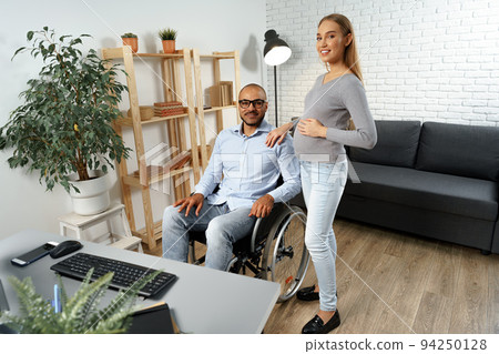 Pregnant woman holding a hand of her disabled husband sitting in a wheelchair Pregnant woman holding a hand of her disabled husband sitting in a wheelchair 94250128