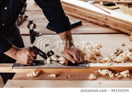 Carpenter's hands planing a plank of wood with a hand plane Carpenter's hands planing a plank of wood with a hand plane 94250204