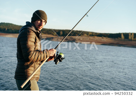 Young fisherman standing on the shore of lake with fishing rod Young fisherman standing on the shore of lake with fishing rod 94250332